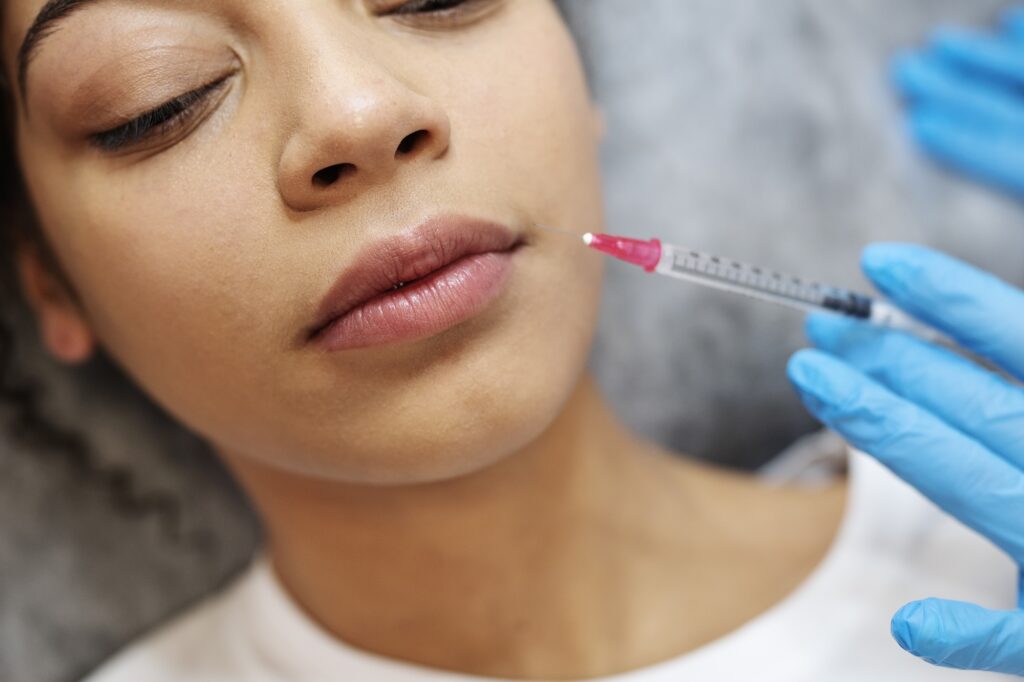 A young woman laying down in a treatment room at Clarity Health & Aesthetics while a provider leans over her, getting ready to inject her lip fillers near Kansas City.